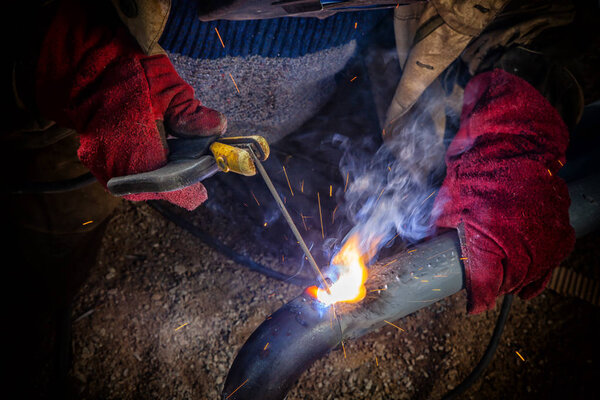 Close up welder weld metal pipe at construction site. A  builder in brown uniform, welding mask and welders leathers, weld metal with a arc welding machine, blue sparks fly to the sides