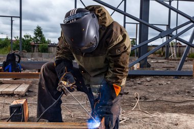 Kaynakçı Kaynak metal boru inşaat sahasında kapatın. Maske ve kaynakçılar deri, kaynak bir Oluşturucu kahverengi üniformalı bir ark kaynak makinesi, iki yanda mavi kıvılcım sinek ile Kaynak metali