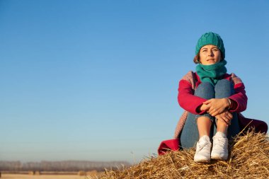 Mavi örgü şapka ve pembe ceket haystack üzerinde oturan genç çekici bayan. Gülümseyen güzel hipster mutlu kadın şık giyim eşyası alan üzerinde. Sonbahar eğilim, kentsel stydent tarzı.