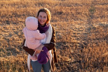 Örgü şapka ve sıcak giysiler ile küçük bebek oyun, güzel bir genç model portresi gün, arka plan haystacks güneşli sonbahar gün tadını çıkarın. Sonbahar sıcak fotoğraf. Kadın gülümsüyor ve göz uzak, neşeli neşeli ruh.