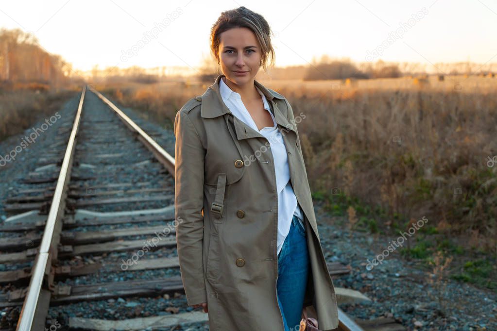 A young woman in a white shirt, beige raincoat and jeans  enjoys nature, walking along the railroad tracks around blue sky. The concept of livestyle and outdoor recreation in autumn