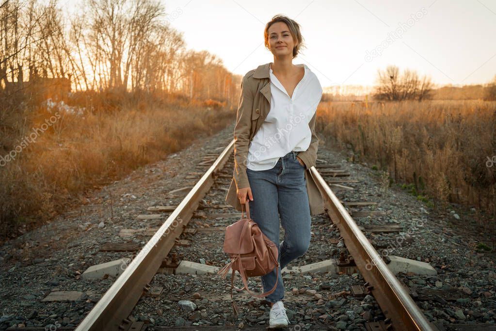 A young woman in a white shirt, beige raincoat and jeans  enjoys nature, walking along the railroad tracks around blue sky. The concept of livestyle and outdoor recreation in autumn