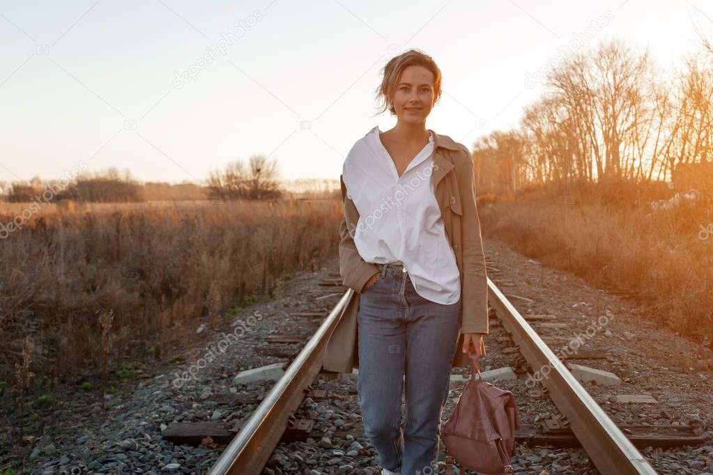 A young woman in a white shirt, beige raincoat and jeans  enjoys nature, walking along the railroad tracks around blue sky. The concept of livestyle and outdoor recreation in autumn