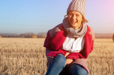 Güzel bir genç model gri örgü şapka ve sıcak giysiler portresi eğlenmene bak, güneşli bir sonbahar günde alanında oturup. Sonbahar sıcak fotoğraf. Gülümseyen kadın ve kamera, neşeli neşeli ruh göz.