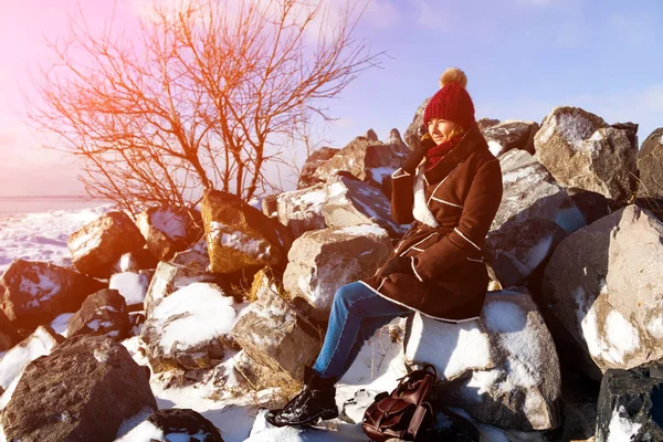 The concept of livestyle  outdoor in winter. A young woman student in a red knitting hat, a brown sheepskin coat sits on a stone rock covered with snow, enjoys winter and looks at the frozen sea on a cold winter day