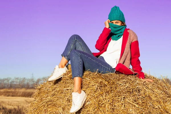 Outdoor atmospheric lifestyle photo of young beautiful lady with brown hair and eyes in pink coat, knitting hat and jeans posing and sitting on hay around  blue sky. Warm autumn.