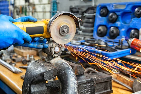 A close-up of a car mechanic using a metal grinder to cut a car silent block in a vise in an auto repair shop, bright flashes flying in different directions, in the background tools for an auto repair shop. Work of auto mechanics.