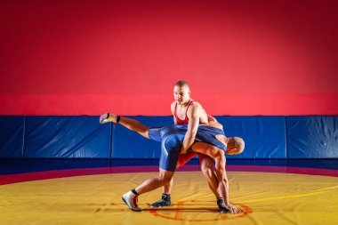 Two strong wrestlers in blue and red wrestling tights are wrestlng and making a suplex wrestling on a yellow wrestling carpet in the gym. Young man doing grapple. clipart