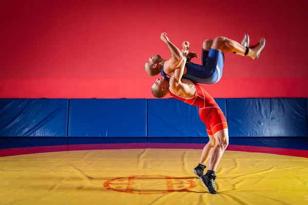 公平摔跤的概念 两名身穿红蓝相间制服的男子摔跤选手在体育馆的一块黄色摔跤地毯上摔跤 — 图库照片