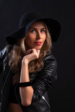 Gülümseyen güzel genç kadın trendy siyah T-shirt, turuncu etek, deri ceket ve siyah şapka ile poz çene üzerinde eller. Üç çeyrek uzunluğu studio siyah arka plan üzerine vurdu.