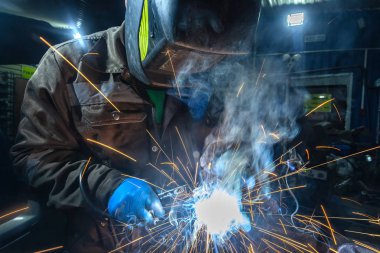 Man welder in welding mask, building uniform and blue protective gloves welds metal car muffler with welding machine in auto repair shop, in the background construction site clipart