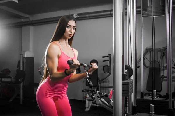 Dark-haired woman athlete in a tracksuit shakes hands biseps on the simulator in the gym. Woman trainer shows how to shake hands for women