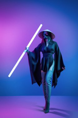 Kozmik cosplay kavram. Çağdaş portre geleneksel Japon siyah kimono, bir Asya şapka ve highboots bir genç atletik kadında bir ışın kılıcı tutan ve neon mavi-pembe arka plan üzerinde poz