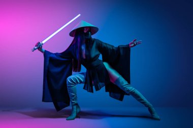 Concept on cosmic cosplay. Contemporary portrait a young athletic woman in traditional Japanese black kimono, an Asian hat and highboots is holding a lightsaber and posing on neon blue-pink background clipart