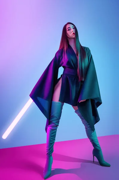 Concept on cosmic cosplay. Contemporary portrait a young athletic woman in traditional Japanese black kimono, an Asian hat and highboots is holding a lightsaber and posing on neon blue-pink background