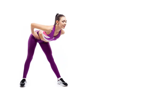 A dark-haired woman coach in a sporty pink short top and gym leggings shows the correct technique of tilting forward for inflating the buttocks on a  white isolated background in studio