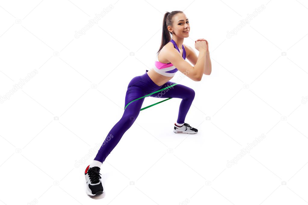 A dark-haired woman coach in a sporty purple short top and gym shows the correct squatting technique with sport fitness rubber bands; the position of a full squat  on a  white isolated background in studio