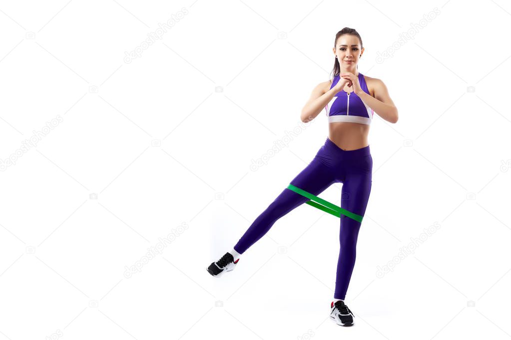 A ypung woman coach in a sporty purple  short top and gym leggings makes lunges  by the feet forward with sport fitness rubber bands,  stretch legs on a  white isolated background in studio 