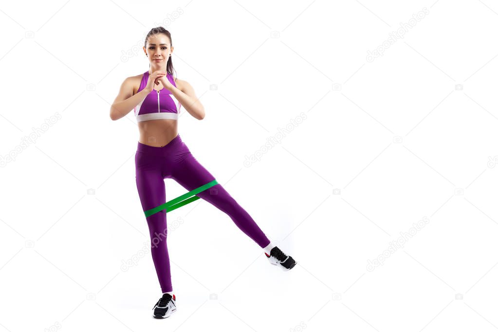A ypung woman coach in a sporty pink   short top and gym leggings makes lunges  by the feet forward with sport fitness rubber bands,  stretch legs on a  white isolated background in studio 
