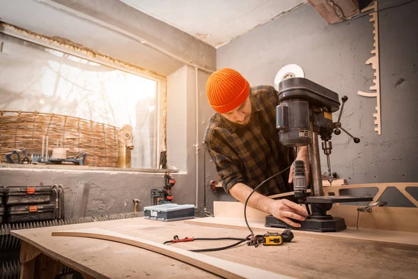 A man with work clothes and a carpenter\'s cap is carving a wooden board on an modern large drilling machine in a light workshop