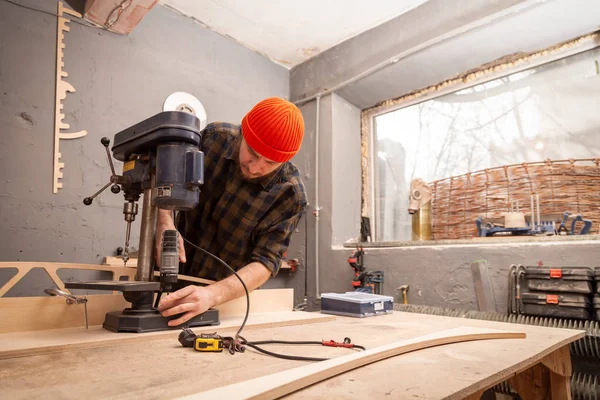 A man with work clothes and a carpenter\'s cap is carving a wooden board on an modern large drilling machine in a light workshop