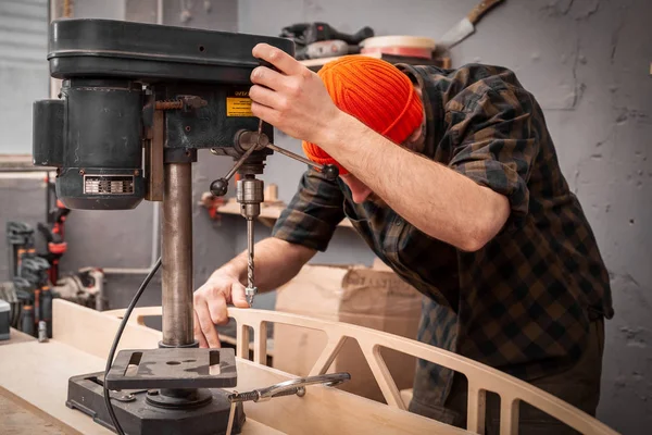 A man with work clothes and a carpenter's hat is carving a wooden board on an  large drilling machine in a light workshop