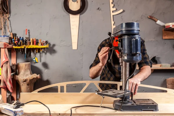 Close up of a man with work clothes and a carpenter\'s cap is carving a wooden board on an modern large drilling machine in a light workshop. Home repair concepts