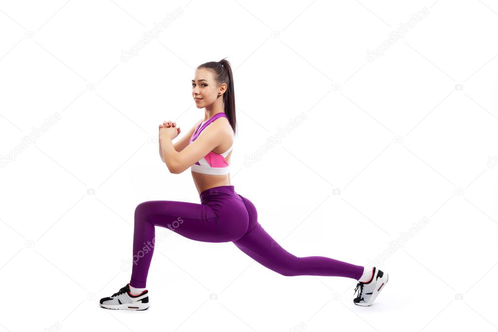 A young  woman coach in a sporty  short top and gym leggings makes lunges  by the feet forward, hands are held out to the side   on a  white isolated background in studio 