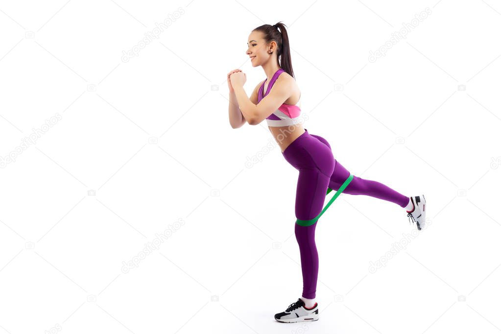 A young  woman coach in a sporty  short top and gym shows the correct makes lunges with sport fitness rubber bands on a  white isolated background in studio