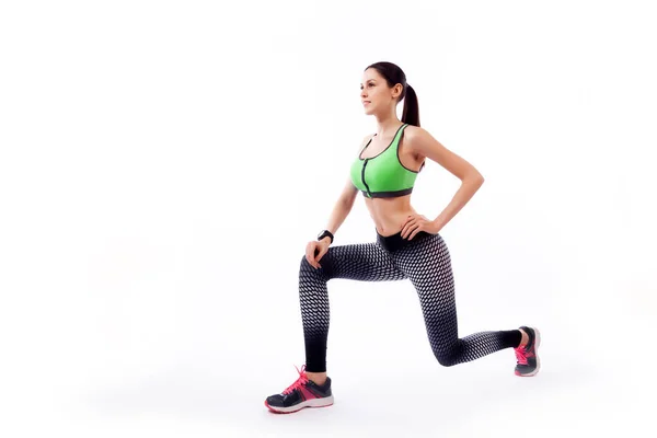 A dark-haired woman coach in a sporty short top and gym leggings makes lunges  by the feet forward, hands are held out to the side   on a  white isolated background in studio