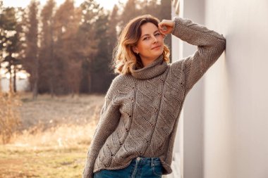 Doğal yün ve kot ten yapılmış kahverengi örgü kazak giymiş genç moda kadın Moda yaşam tarzı portre gülerek, gülümseyerek, poz ve sokakta beyaz bir duvara karşı . Neşeli kadın portresi