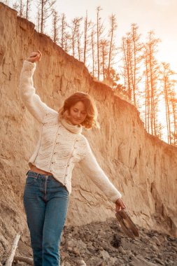 Doğal yün ve kottan yapılmış kahverengi örgü kazak giymiş genç moda kadın Moda yaşam tarzı portre gülerek, gülümseyerek, plaj poz; ağaçlar ile arka planda kumlu yamaç. Neşeli kadın portresi