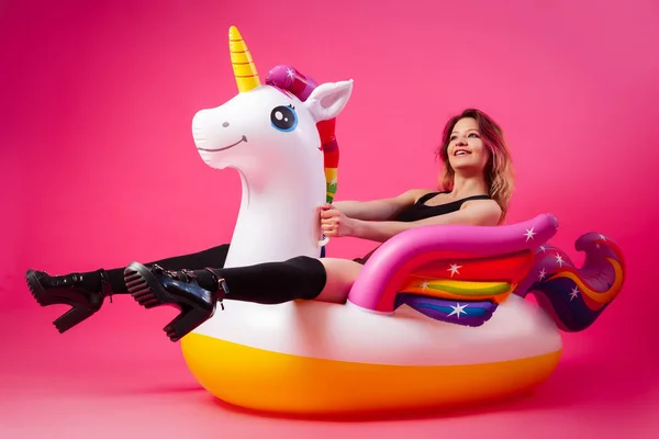 Concept summer mood, relaxation and beauty.Modern portrait of a young woman in a yellow swimsuit, bright socks and sandals resting on an inflatable unicorn mattress on an isolated pink-blue background
