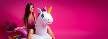 Konsept yaz ruh hali, gevşeme ve güzellik. Bir şort içinde genç bir kadının modern portre, üst ve sandalet izole pembe arka planda şişme tek boynuzlu yatak üzerinde dinlenme