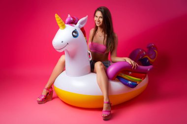 Konsept yaz ruh hali, gevşeme ve güzellik. Bir şort içinde genç bir kadının modern portre, üst ve sandalet izole pembe arka planda şişme tek boynuzlu yatak üzerinde dinlenme