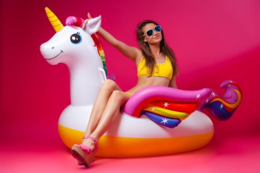 Yaz moda kız, pembe ve neşeli ruh hali. Güneş gözlüğü güzel ince kadın, sarı mayo oturur, eğlenceli ve pembe izole arka planda beyaz şişme Unicorn yaşam sahiptir