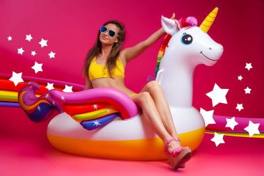 Yaz moda kız, pembe ve neşeli ruh hali. Güneş gözlüğü güzel ince kadın, sarı mayo oturur, eğlenceli ve pembe izole arka planda beyaz şişme Unicorn yaşam sahiptir