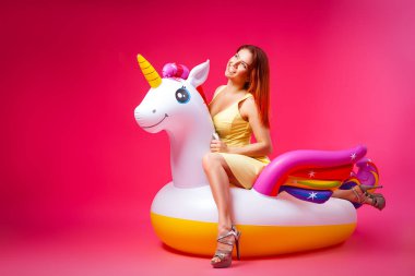 Konsept yaz ruh hali, rahatlama ve güzellik. Izole pembe bir arka plan üzerinde şişme tek boynuzlu at şilte üzerinde dinlenmiş sarı elbiseli genç bir kadının modern portresi