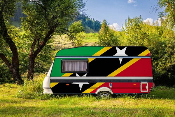 A car trailer, a motor home, painted in the national flag of  Saint Kitts and Nevis stands parked in a mountainous. The concept of road transport, trade, export and import between countries. Travel by car