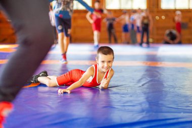 Altay, Rusya 16 Ağustos 2019: Kırmızı güreş taytlı küçük çocuk spor salonunda mavi güreş halısında bir spor kampında spor egzersizi yapıyor ve neşeyle gülümsüyor. Açık eğitim ve yaz spor kampları 