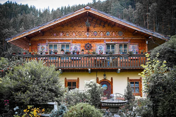 Authentic wooden two-story house with a fountain with painted walls, shutters and windows stands, surrounded by a lot of trees, bushes and various flowers and plants, in the background mountains and forest