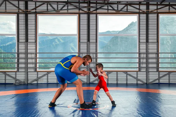 A little wrestler boy in a sports tights wrestles with an adult male wrestler on a wrestling carpet in the gym. The concept of child power and martial arts training. Teaching children Greco-Roman wrestling