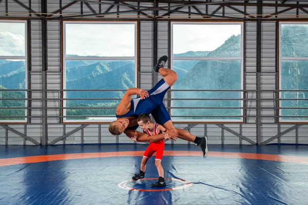 スポーツタイツのレスラー少年は ジムのレスリングカーペットの上で大人の男性レスラーと格闘します 子どもの力と武道の訓練の概念 子供たちにグレコローマレスリングを教える — ストック写真