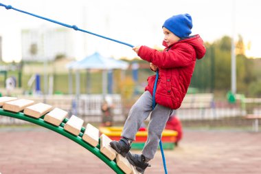 Sıcak kırmızı ceket ve şapka küçük bir komik çocuk bir şehir parkı bir oyun alanında bir ip kullanarak ahşap bir slayt tırmanıyor. Çocuk eğlence konsepti