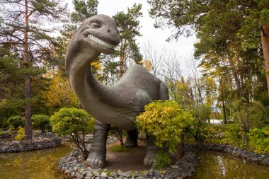 Büyük gri yetişkin bir brachiosaurus çerçeveye doğru bakar ve sıcak bir sonbahar gününde küçük ağaçların ve çalıların yanında nehrin taş kıyısında durur. Dinozor dünyası