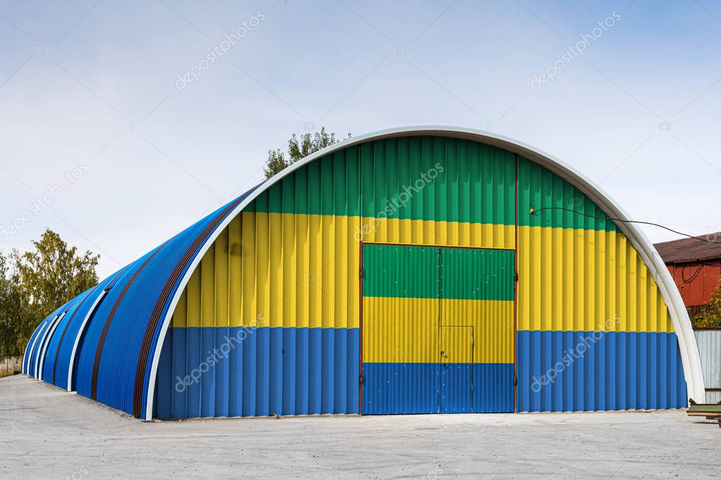 Close-up of the national flag of Gabon painted on the metal wall of a large warehouse the closed territory against blue sky. The concept of storage of goods, entry to a closed area, logistics