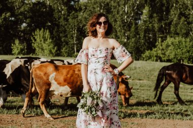 Açık hava atmosferik yaşam tarzı portresi pembe elbiseli güzel, koyu renk saçlı bir kadının bir inek sürüsünün arka planında duruyor. Kız kendini seyahate çağırıyor.