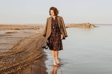 Koyu renk saçlı bir kadın gülümser, sahil boyunca yürür ve yaz gününde parlak güneşin tadını çıkarır. Denizde yaz tatili ve yaşam tarzı kavramı