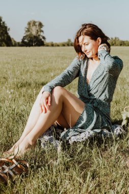 Açık hava moda portresi açık mavi elbiseli yeşil çimenlerde oturan genç bir kadın. Yaz kıyafetleri ve aksesuarlar..