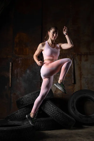 A dark-haired woman coach in a sporty  pink  short top and gym leggings doing exercise to warm up the legs, alternate knee flexion  on a  in an old garage, against the background of tires. Conceptual Sports Photo